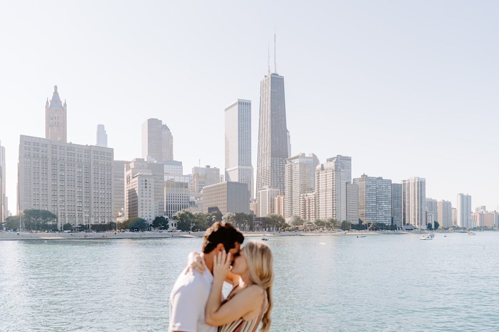 Fiancés snuggle in tight and hug on the lake front in Chicago during their engagement session at Milton Lee Olive Park with the Chicago skyline in focus