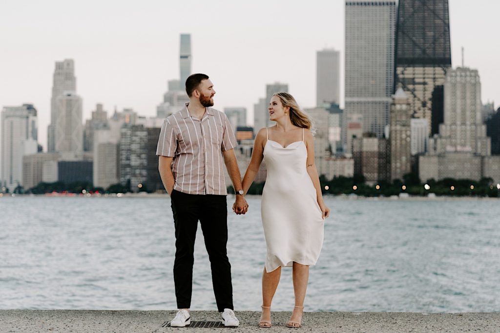 An engaged couple stands next to each other at their engagement session on North Avenue Beach.  The Bride is wearing a silk dress and the groom is wearing a pink shirt with white stripes.