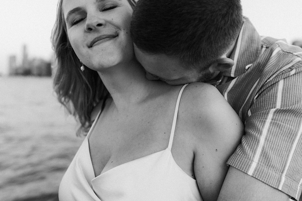 A black and white photo of a future groom kisses his future bride on the neck during their beach engagement session overlooking Lake Michigan