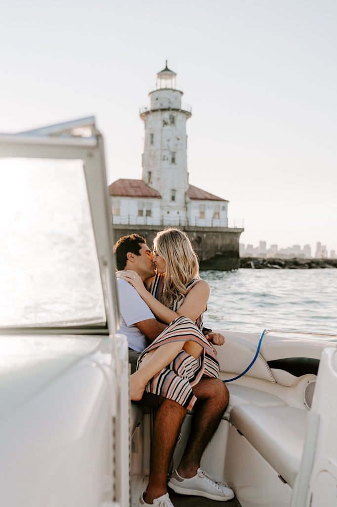 Fiancés kiss and hold each other tight on a boat in Chicago with a lighthouse in the background during their engagement session