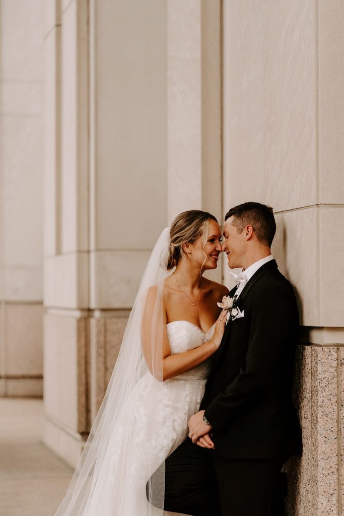 A bride and groom press their noses together standing against a marble wall outside their wedding venue in the Chicago suburbs.