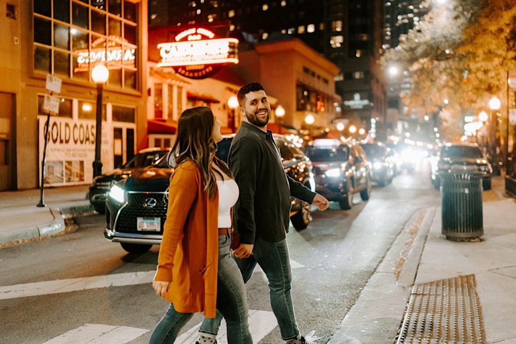 An engaged couple crosses the street at night in Gold Coast Chicago while smiling at each other and holding hands.