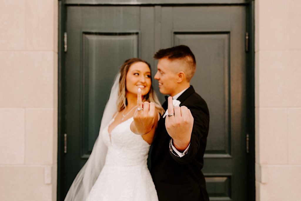 Bride and groom on their wedding day holding up their ring fingers towards the camera while looking at one another.  Their hands and ring fingers are in focussed.