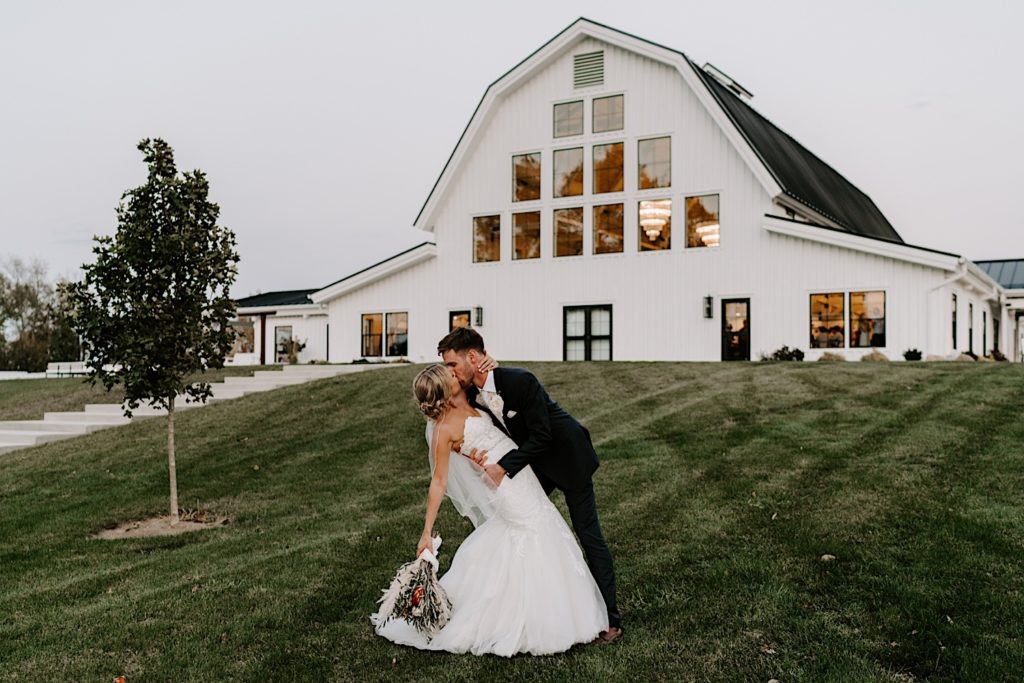 A bride and groom kiss while the bride dips backward with the support of her groom while standing in front of their wedding venue that is a white barn with a lot of windows.