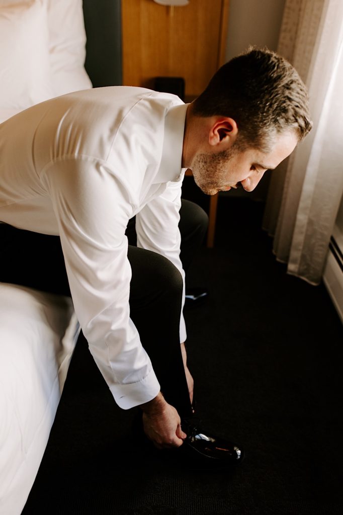 A groom puts on his shoes before his wedding day while sitting on the side of a bed in a hotel room.