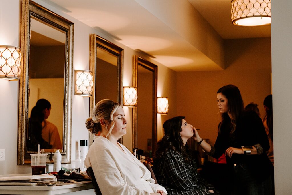 A bride sits with her eyes closed next to a bridesmaid getting her makeup done for the wedding day.