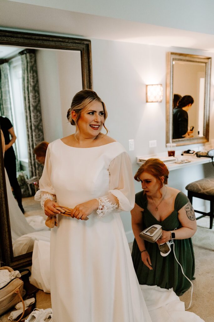 A bride standing in her wedding dress while smiling over her shoulder. Behind her a bridesmaid is crouched looking for any wrinkles or creases to iron out of the dress.