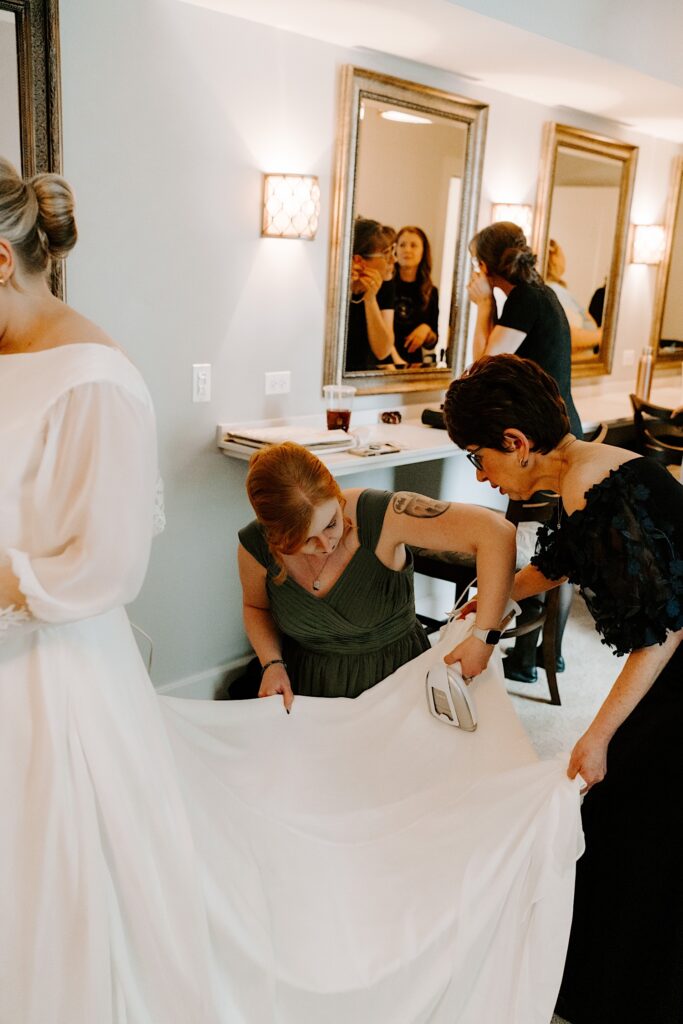 A bridesmaid irons out a wrinkle of a wedding dress being worn by the bride. The mother of the bride is helping hold the dress at the correct angle.