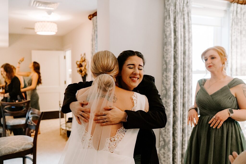 A woman smiles as she hugs a bride wearing her wedding dress while a bridesmaid stands to the left of them. The bride's back is turned to the camera.