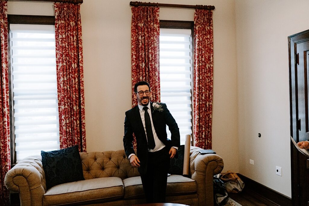 A groom wearing his wedding suit stands in front of a couch and smiles at something out of view .