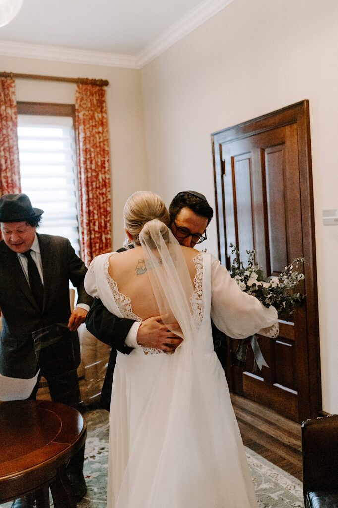 A bride and groom embrace while in their wedding attire after seeing one another for the first time on their wedding day. A man stands off to the right side of them.