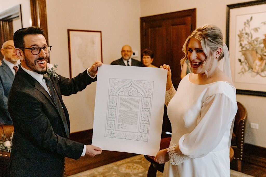 A bride and groom in their wedding attire smile at the camera while holding their signed wedding documents between them.