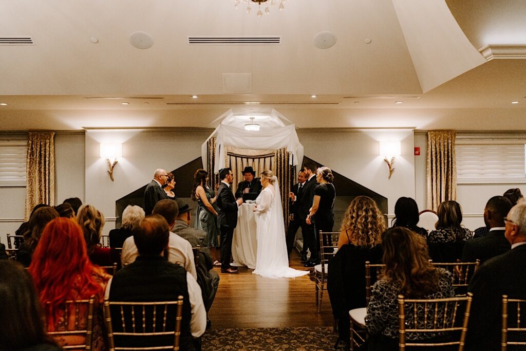 A bride and groom look at one another while the Rabbi gives a speech at their wedding ceremony in Barrington's White House while guests watch