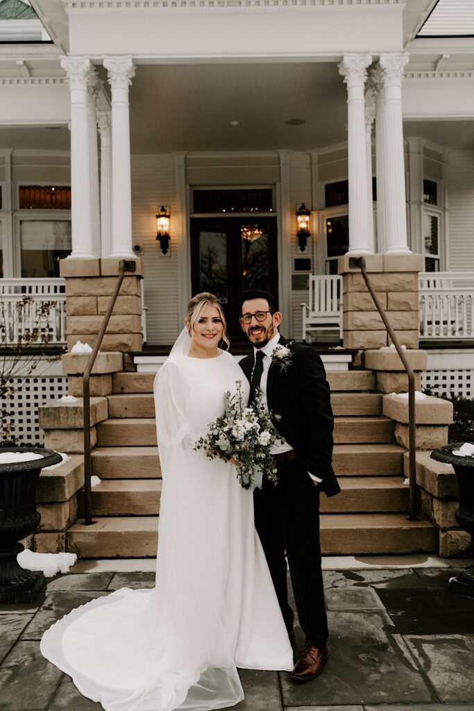A bride and groom stand next to one another and smile outside a building with snow on the stairs