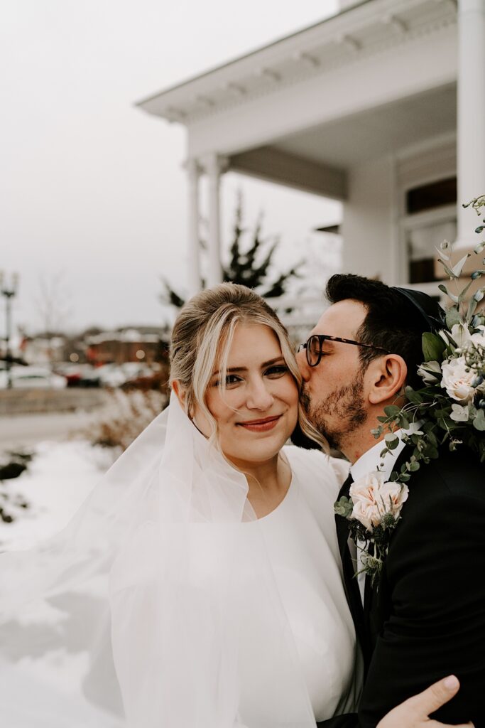 A groom kisses his bride on the cheek while she looks at the camera, they're outside and there is snow on the ground.