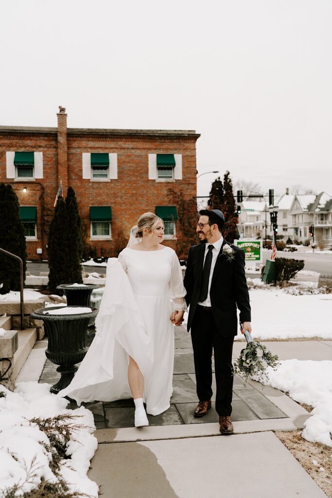 A bride and groom walk hand in hand towards the camera, they're outside and there is snow on the ground