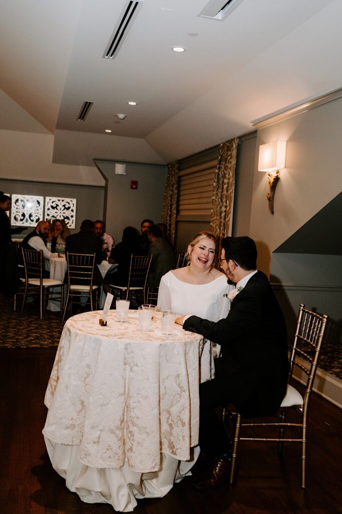 A bride and groom sit and laugh together at their table for their wedding reception.