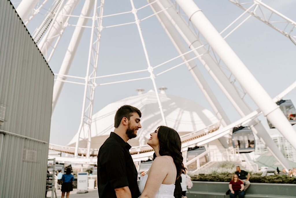 A couple smile at one another underneath the Ferris Wheel at Navy Pier in Chicago during their engagement session