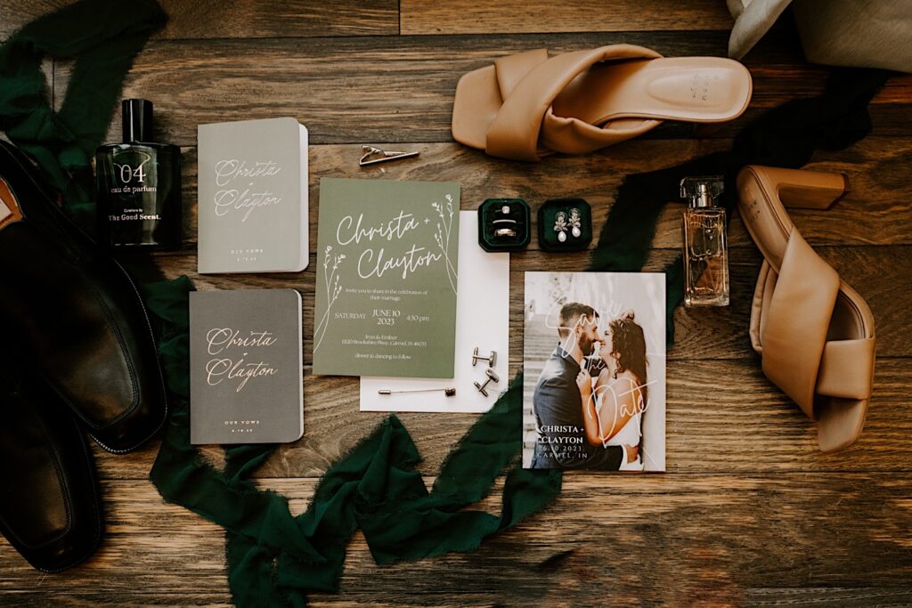 A wedding day flat lay consisting of invites, RSVP's, perfume, cologne, shoes, rights, and a green ribbon on a wood floor