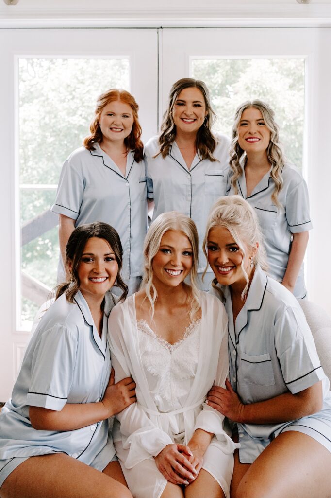 A bride and her 5 bridesmaids all smile at the camera while wearing their robes before getting ready for a wedding