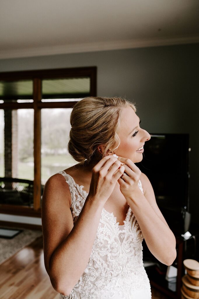A bride in a house puts on her earrings while looking out a window as she gets ready for her wedding day