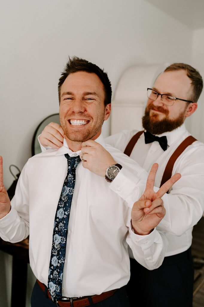 A groom smiles and puts peace signs up as a groomsman stands behind him and adjusts his tie as they get ready for a wedding