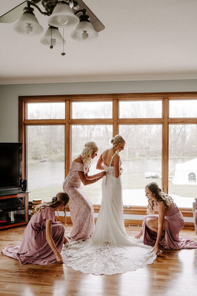 A bride stands in front of a window in her wedding dress as her mother and two bridesmaids help button it up and lay out the wrinkles