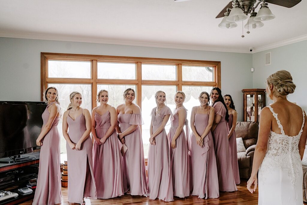 Bridesmaids turn and smile as they see the bride standing in front of them in her wedding dress for the first time