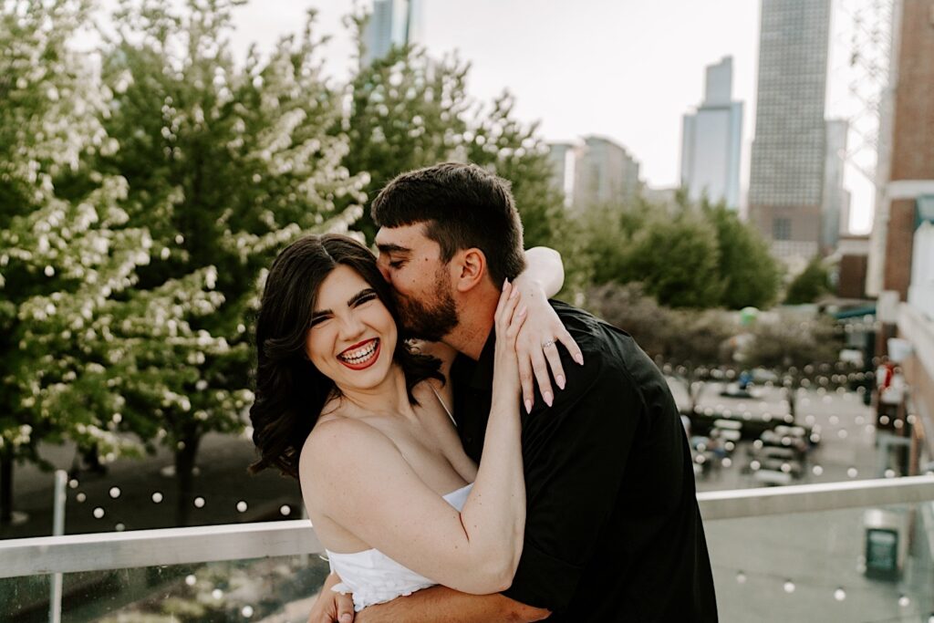 During an engagement session a woman smiles at the camera as a man kisses her cheek while on a balcony at Navy Pier looking out over the street and the Chicago skyline