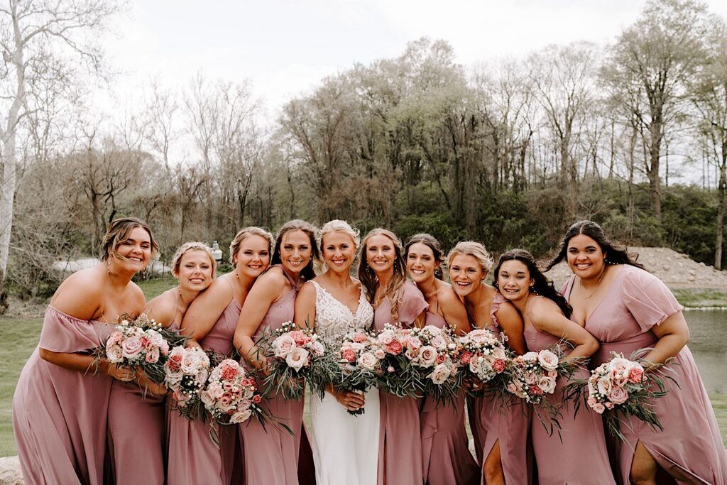 A bride and her bridesmaids who are on either side of her all smile at the camera while standing in the backyard where the tent wedding reception will take place