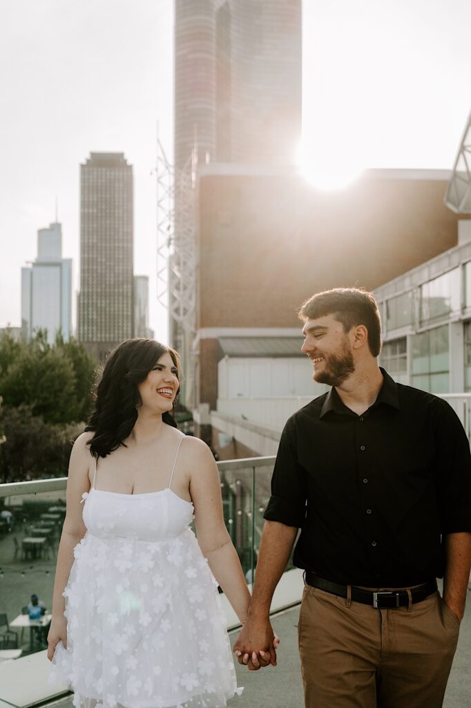 A couple walk towards the camera while holding hands and smiling at one another while the sun sets behind them on the Chicago skyline