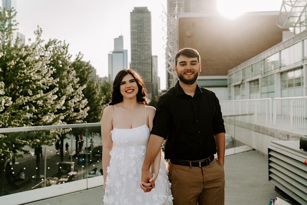 A couple smile and pose during their engagement session at Navy Pier in Chicago, the sun is setting between the buildings behind them