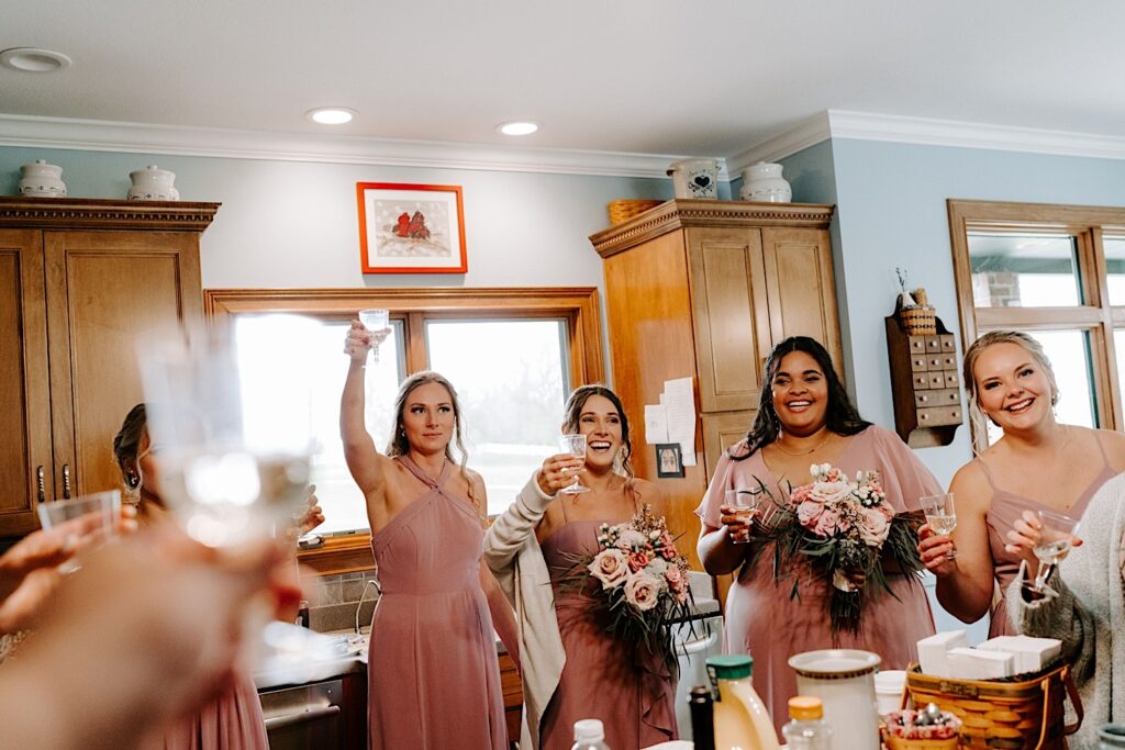 Bridesmaids all stand in a kitchen each with a glass of champagne raised to the sky for a toast before a wedding
