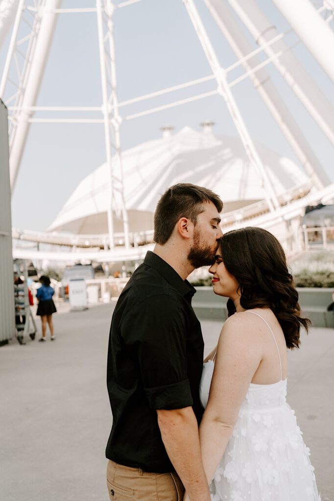 A man kisses a woman on the forehead as she smiles, they're at Navy Pier in Chicago with the Ferris Wheel behind them