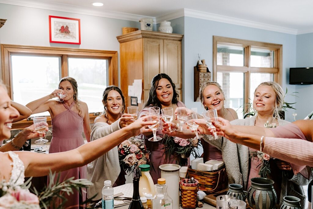 Bridesmaids in a kitchen all clink their champagne glasses together and smile during a speech before a wedding day