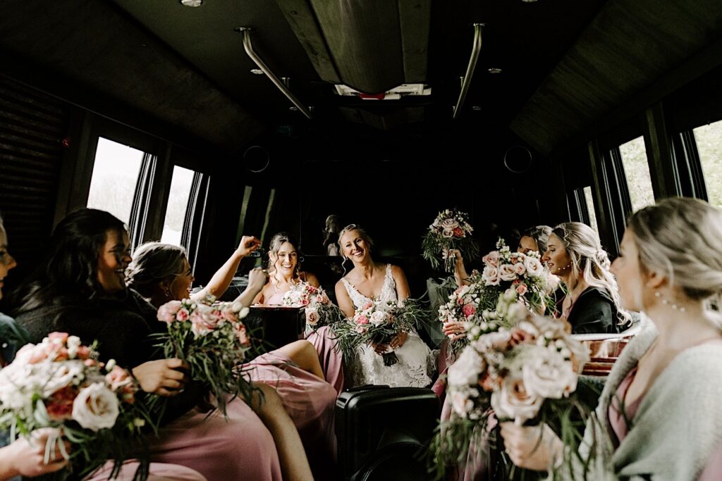 A bride rides in a limo and smiles at the camera while her bridesmaids all sit with her, each of them holding a bouquet of flowers