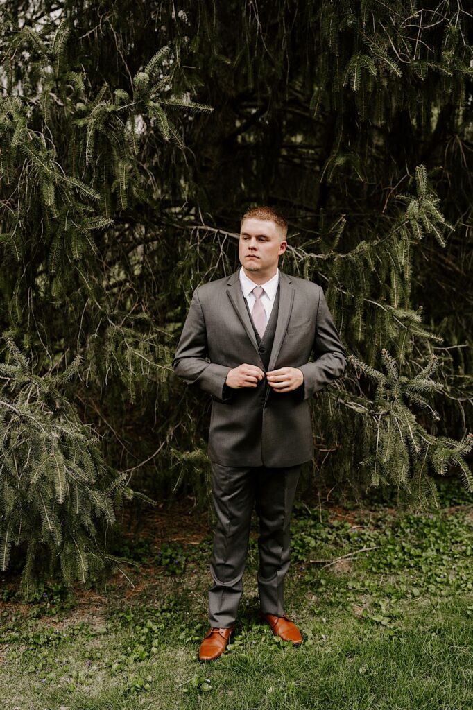A groom stands in front of a pine tree and looks left while buttoning up his suit coat