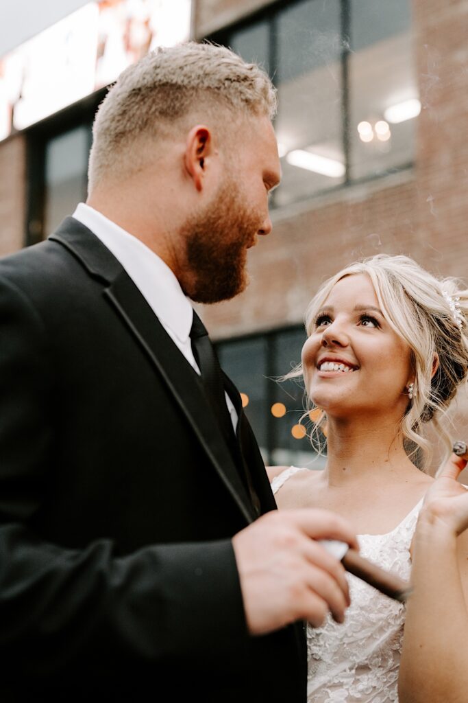 A bride and groom smile at one another while standing outside of a brick building, each of them is holding a lit cigar