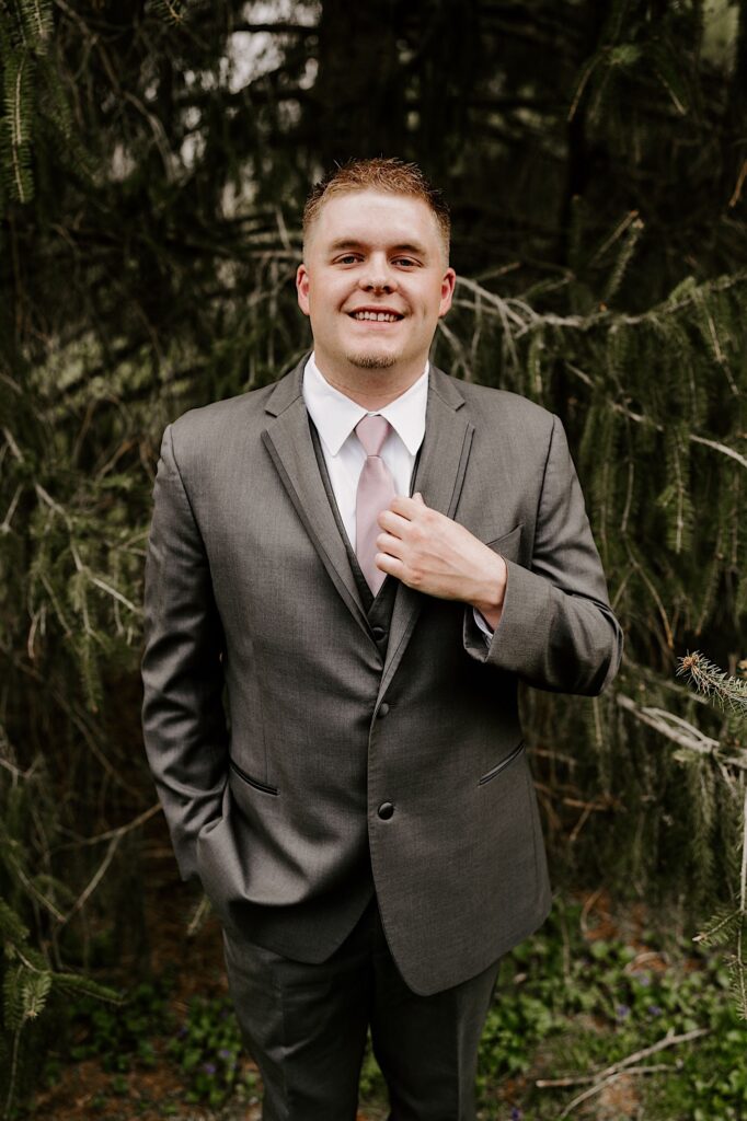 A groom adjusts his suit coat while smiling at the camera and standing in front of a pine tree