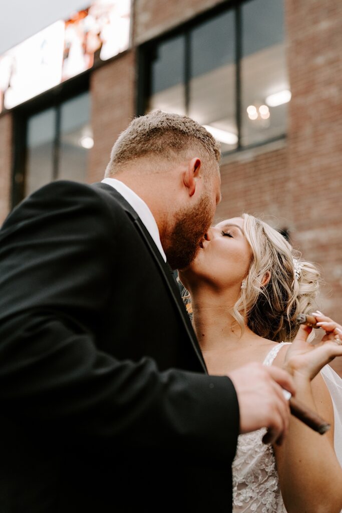 A bride and groom kiss one another while standing outside of a brick building, each of them is holding a lit cigar