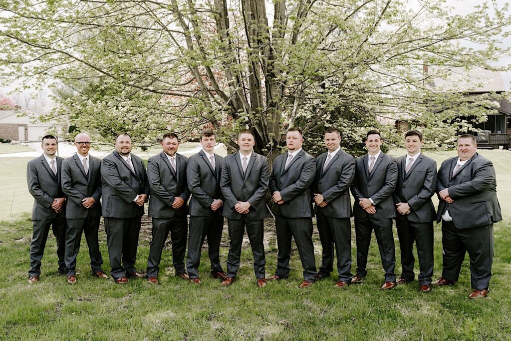 A groom smiles at the camera while his groomsmen stand on either side of him, all of them stand in front of a large tree in the yard of a house