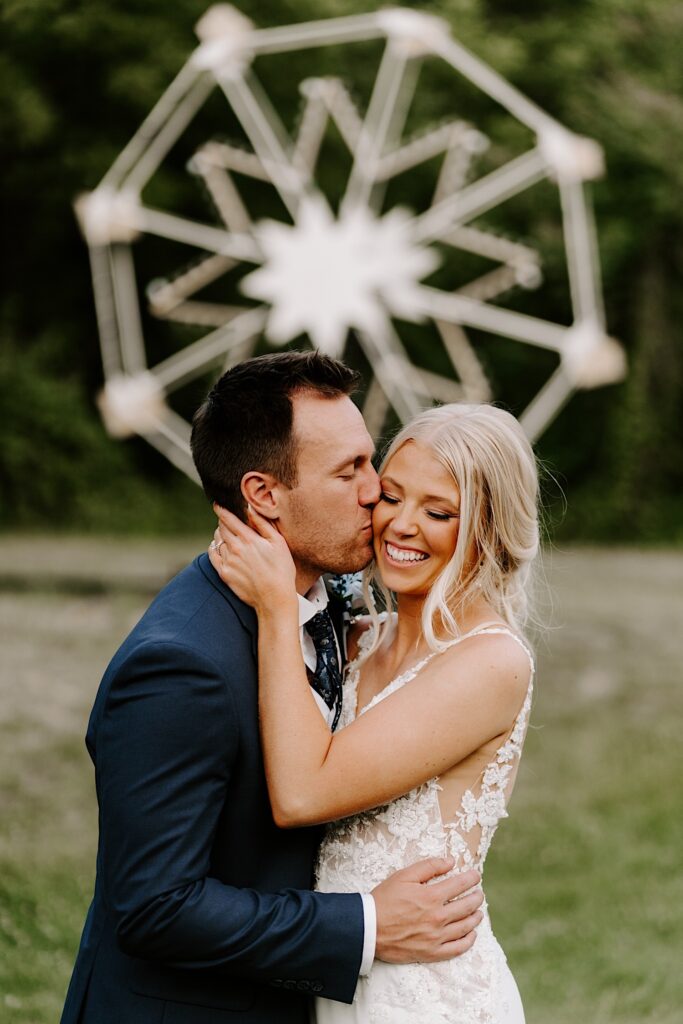 A groom kisses his bride on the cheek as she smiles while they stand in a field with a Ferris Wheel in the background