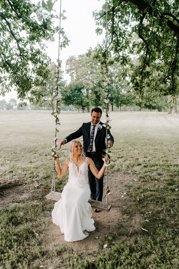 A bride sits on a swing with decorating it in the middle of a park as her groom stands behind her about to push the swing