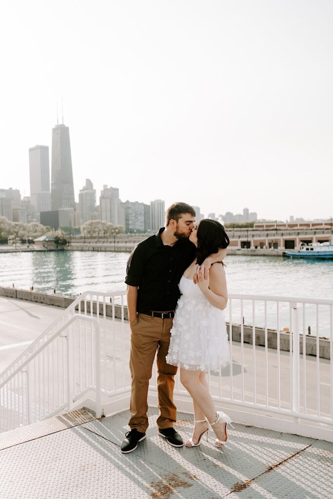 A couple kiss while standing next to a railing on Navy Pier in Chicago with the Chicago skyline and Lake Michigan in the background