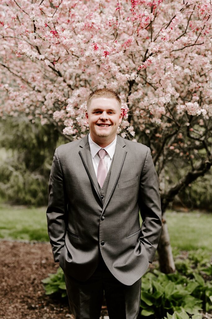 A groom smiles at the camera with his hands in his pockets while standing in front of a cherry blossom tree