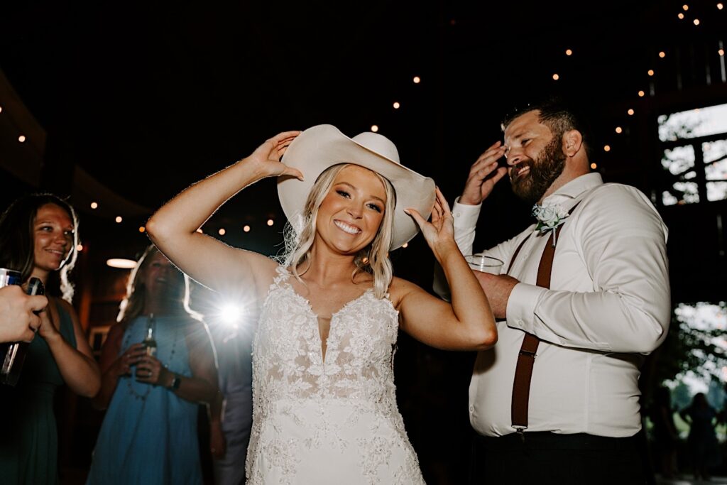 A bride smiles at the camera while wearing a cowboy hat as the groom looks on from behind her and smiles during their indoor wedding reception, photographed by a Chicago wedding photographer