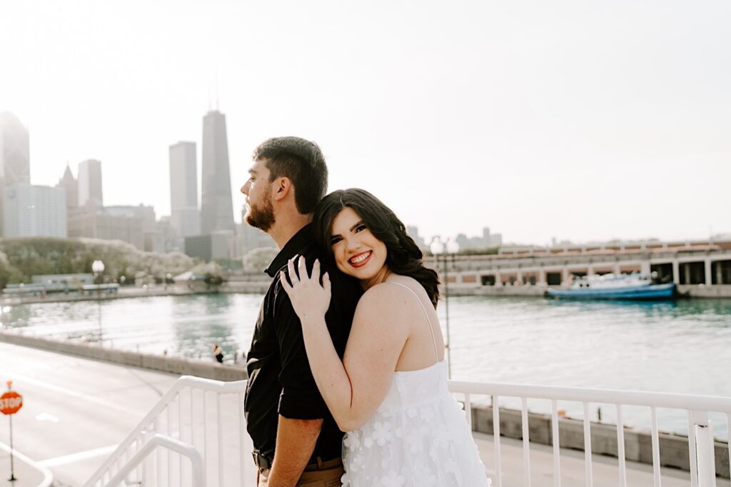 During their engagement session at Navy Pier in Chicago, a man looks to the left while a woman hugs him from behind and smiles at the camera with the Chicago skyline and Lake Michigan in the background