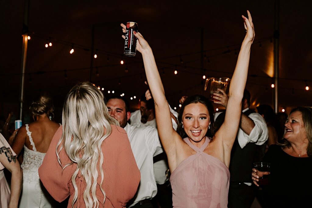 A bridesmaid smiles at the camera and throws her hands in the air, one of them holding a Truly seltzer that she spilled on her dress during an indoor wedding reception, photographed by a Chicago wedding photographer