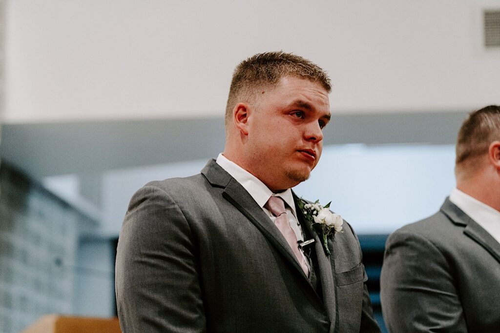 A groom stands in a church during his wedding ceremony and tears up while looking down the aisle and sees his bride for the first time