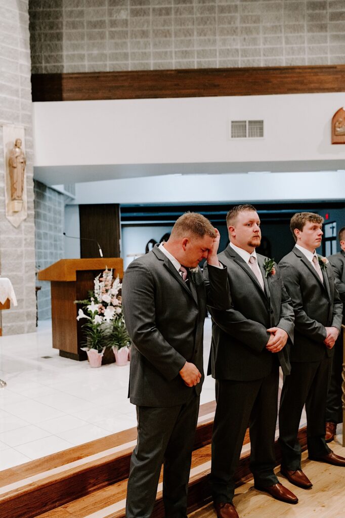 A groom wipes tears from his eyes while standing in a church next to his groomsmen after seeing his bride for the first time